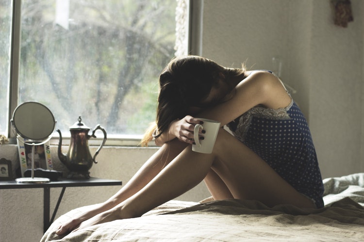 Woman bending over on a bed, her head between her knees as if crying. She is holding a while mug and there is a window behind her with greenery.