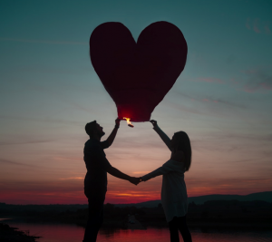 a couple holding a heart shaped lantern balloon against a pink and blue sunset